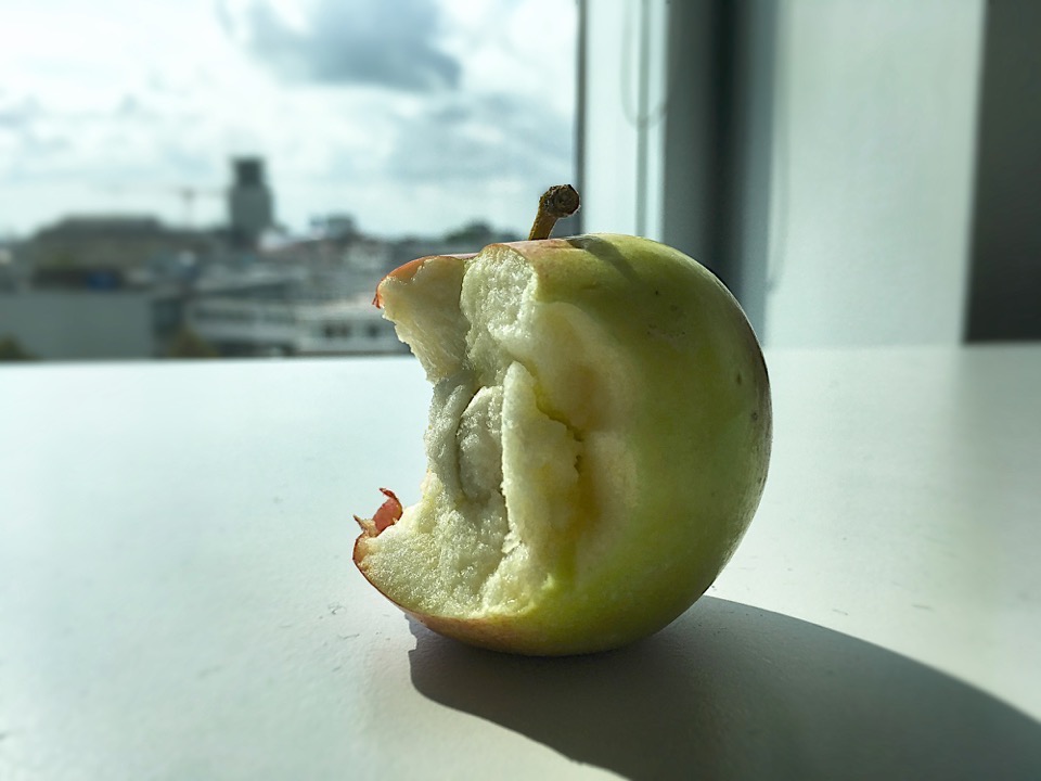 Ein angebissener Apfel liegt auf einem Tisch, im Hintergrund eine verschwommene Stadtsilhouette